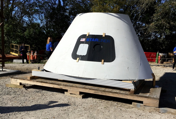 A mock-up of the crew capsule for the reusable CST-100 Starliner, resting under the real Crew Access Arm being built and tested near Kennedy Space Center.